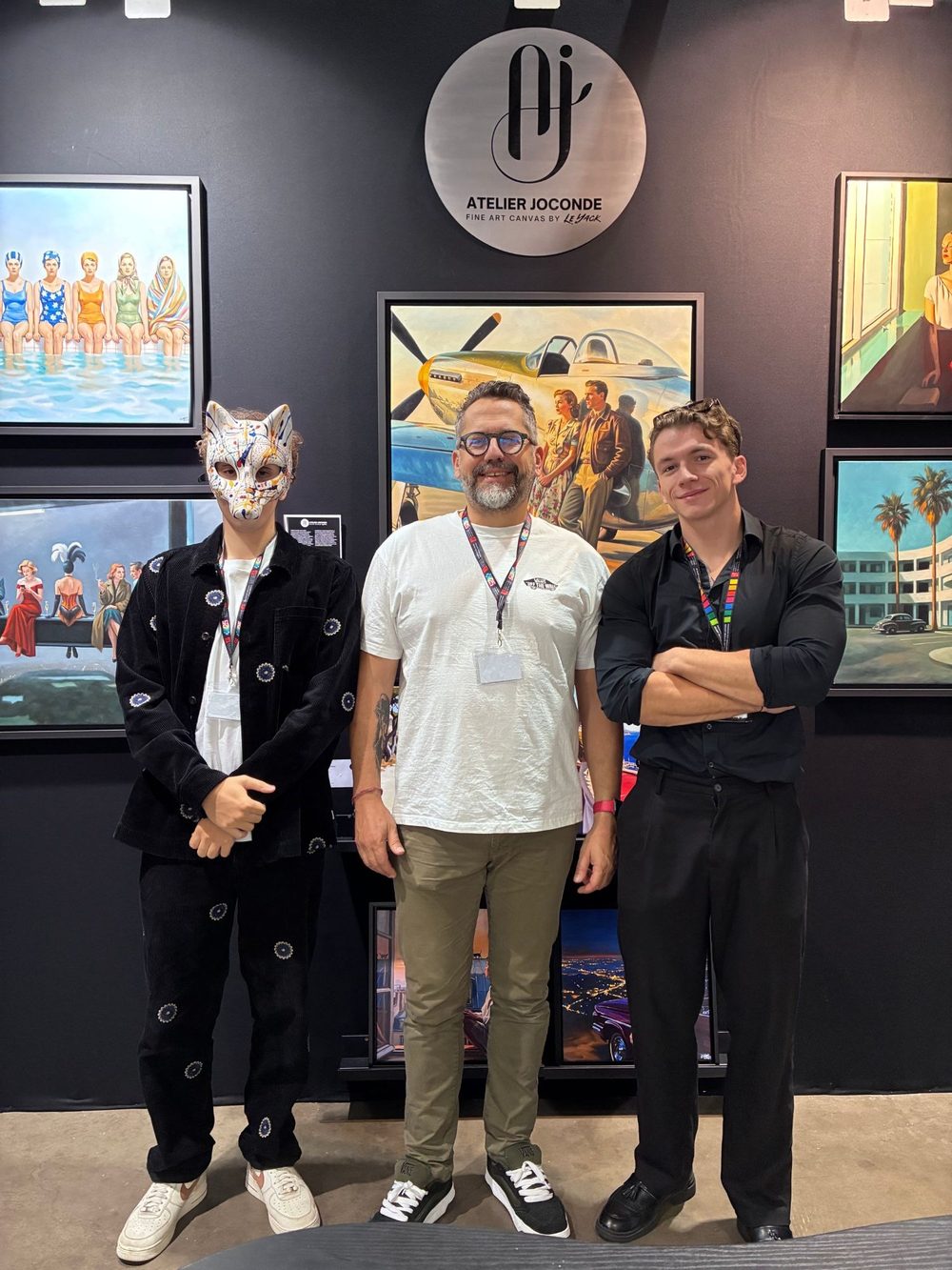 On the Atelier Joconde stand at the art3f fair in Luxembourg, Neo Pablo, masked as a white fox with coloured drips, Le Yack in a white T-shirt and William San in a black shirt, arms crossed, in front of Le Yack’s paintings and the studio’s round sign.