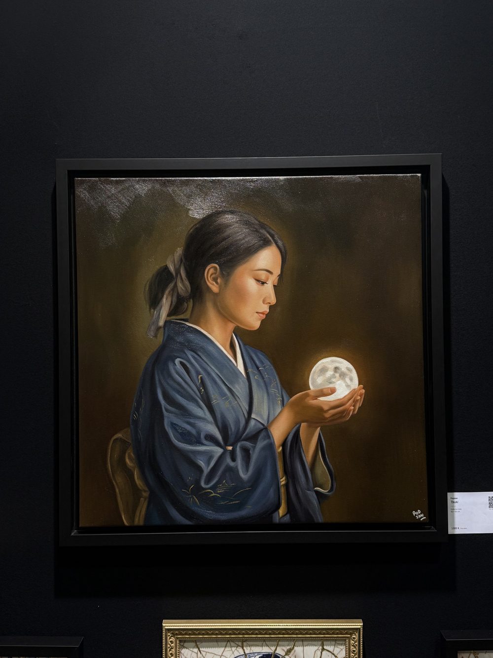 Tsuki, in its black floater frame, hanging on the dark wall of the stand, with its label on the right.
