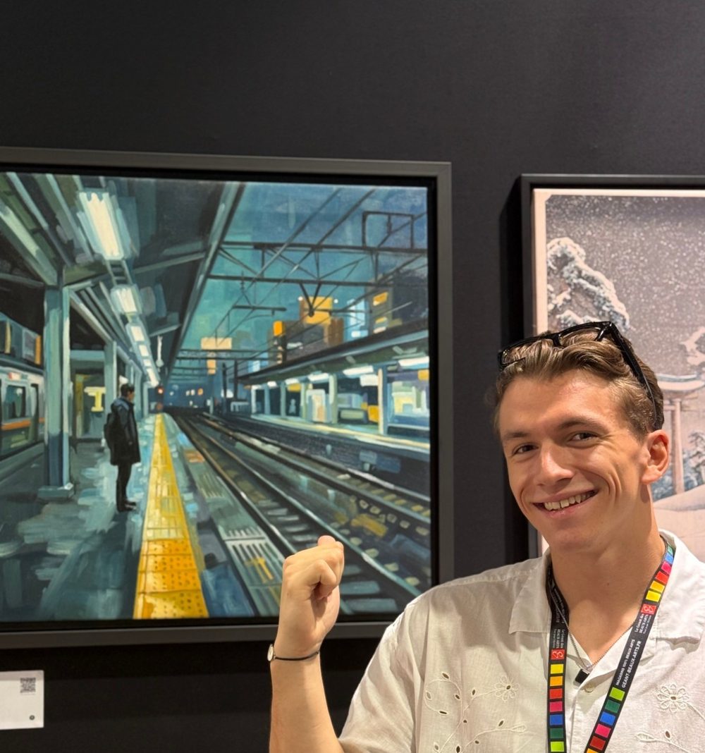 William San, in a white shirt with an Atelier Joconde badge, smiles and raises his fist in front of Shūden, a station platform at night with its yellow line.