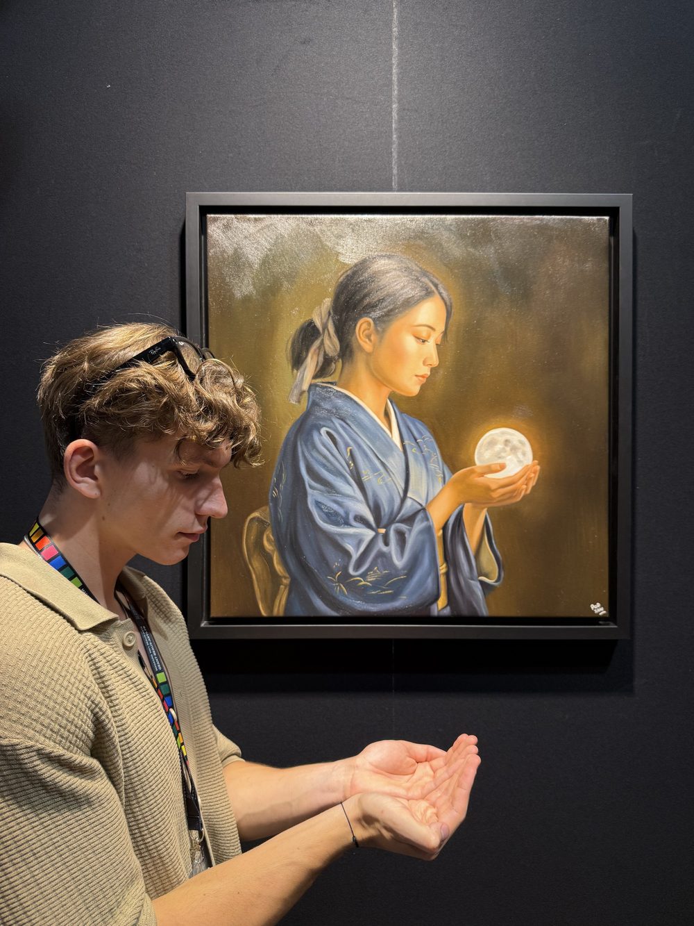 William San, in a beige polo shirt, cups his hands as if holding a moon, beneath Tsuki, a woman in a blue kimono holding a full moon, hanging on the black wall of the stand.