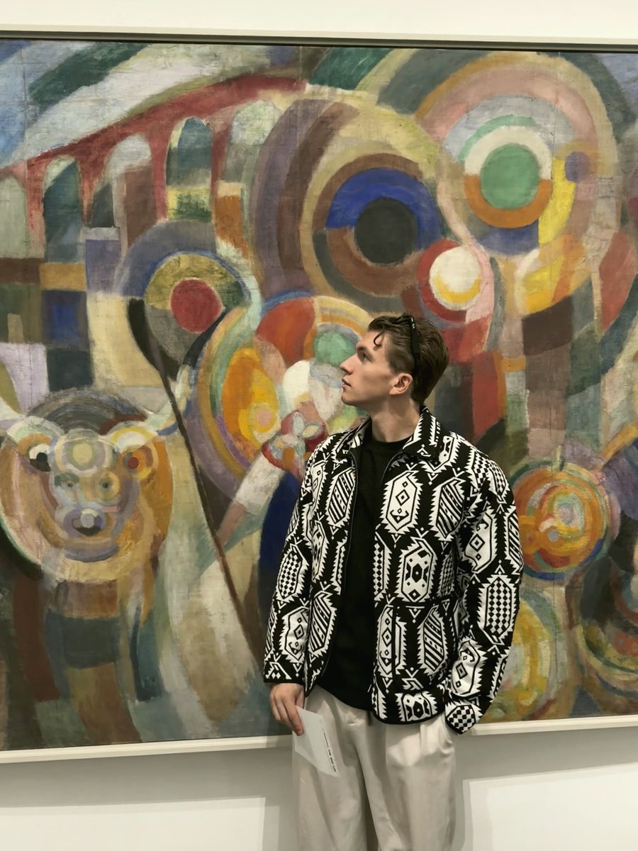 William San in profile, in a patterned black and white jacket, looks at a large abstract canvas of coloured circles in a museum gallery.