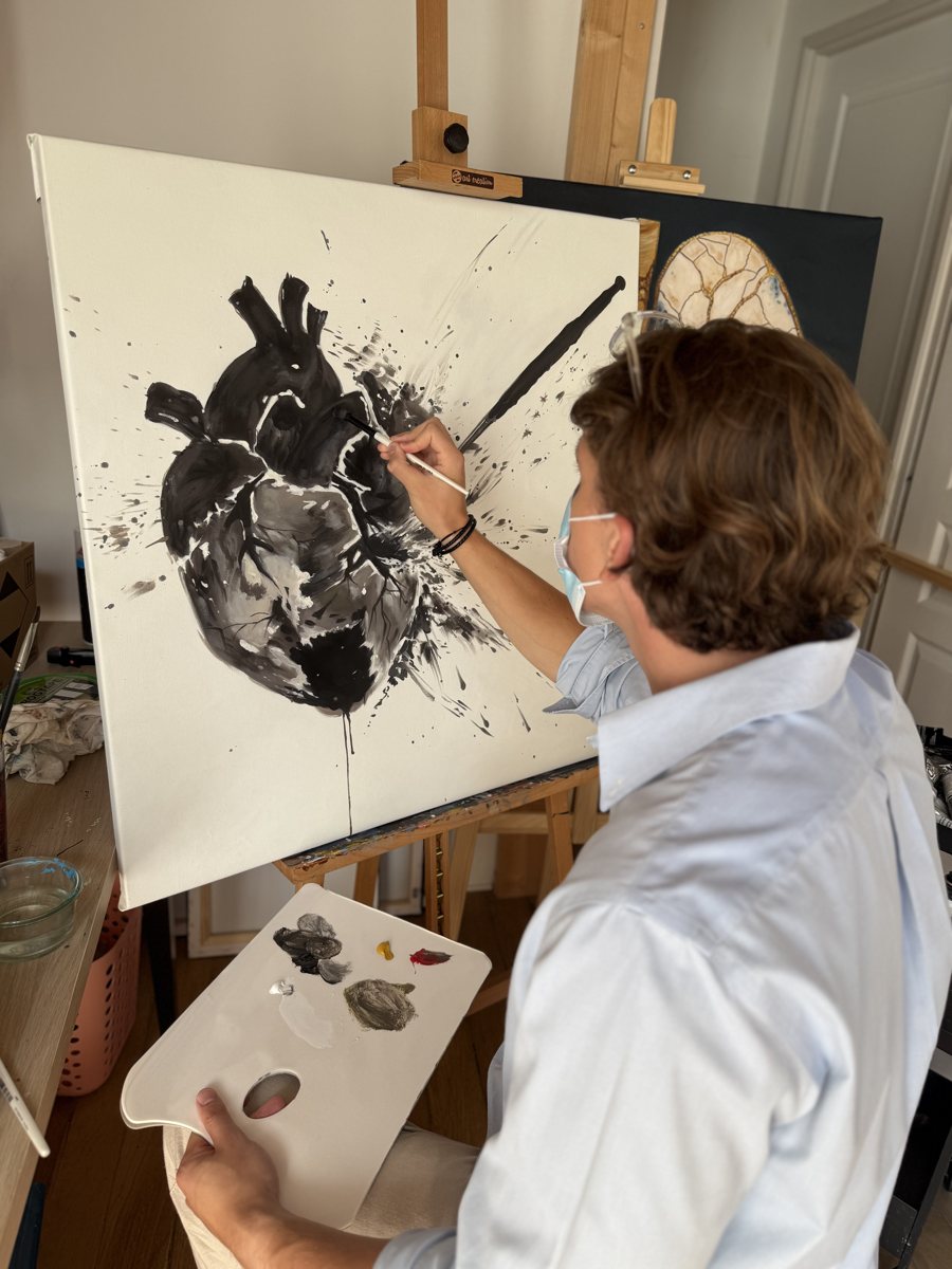 Vu de dos, pinceau et palette en main, William San peint au chevalet Heart Attack, un cœur noir aux éclaboussures d’encre sur une toile blanche.