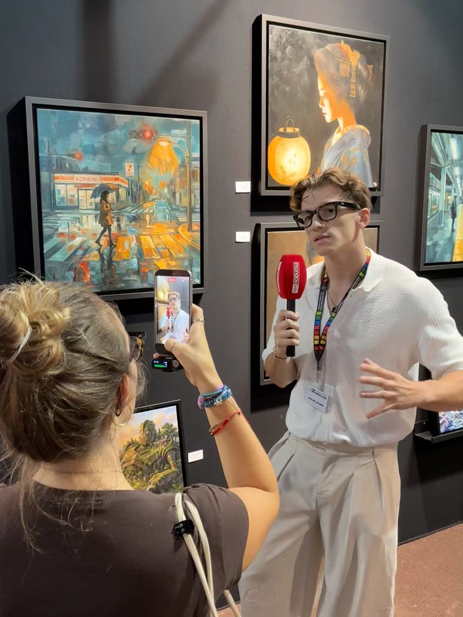 William San, micro rouge en main, répond à une interview sur son stand, devant ses toiles Popsan ; au premier plan, une femme le filme avec son téléphone.